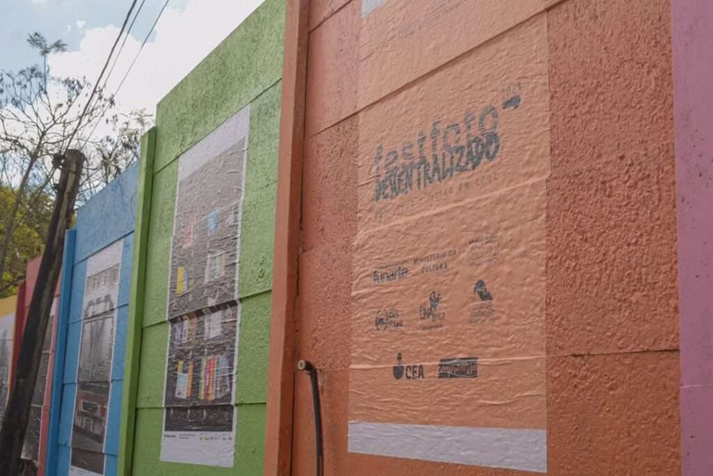 Fotografia de um muro colorido com painéis pintados em tons de azul, verde, laranja e rosa. Neles estão coladas imagens fotográficas e, em destaque, um grande cartaz laranja com o logotipo do "FestFoto Descentralizado 2024" e os dizeres “artistas em movimento cruzado”. Abaixo do logotipo, aparecem logomarcas de apoiadores e patrocinadores, incluindo Funarte, Ministério da Cultura, Banrisul, Fundação Ecarta e outros. À esquerda do cartaz, vê-se parte de uma exposição de fotografias urbanas coladas em sequência sobre o muro. A cena ocorre em ambiente externo, com árvores e fiação elétrica visíveis ao fundo.