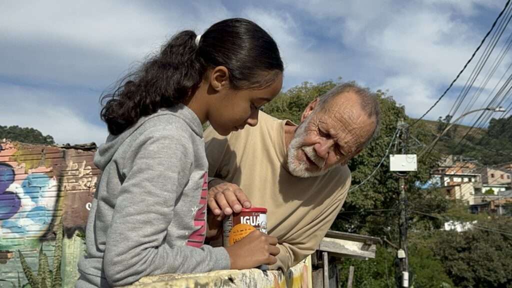 Imagem mostra uma menina e um homem idoso encostados em um muro de concreto com grafites coloridos. A menina, de cabelos escuros presos em rabo de cavalo, veste um moletom cinza e segura uma lata de café. O homem, de barba branca e camisa bege, olha atentamente para a lata enquanto conversa com a menina. Ao fundo, vê-se postes com fios elétricos e casas em uma região de morro, sob um céu azul com nuvens brancas.