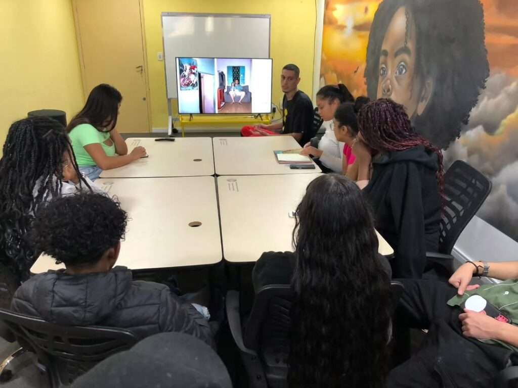 Em uma sala de paredes amarelas e com uma grande pintura artística de uma menina negra com cabelo afro ao fundo, um grupo de adolescentes está reunido em torno de uma mesa retangular formada por quatro mesas brancas unidas. Ao fundo, uma televisão exibe imagens de uma jovem sentada em um quarto. Os jovens assistem atentamente, alguns com cadernos ou celulares à mão. A atmosfera é de atenção e concentração, sugerindo uma atividade educativa ou de formação.
