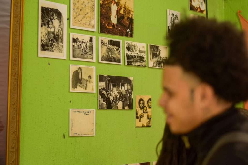 Fotografia mostra uma parede verde decorada com várias fotografias antigas em preto e branco e em tons sépia, organizadas em fileiras. As imagens retratam cenas de cotidiano, reuniões familiares e momentos históricos de comunidades negras. Em primeiro plano, desfocado, está um jovem com cabelo crespo volumoso e lateral raspada, que sorri levemente enquanto observa a exposição. O clima da cena é intimista e afetuoso, sugerindo um ambiente de memória e valorização da ancestralidade.