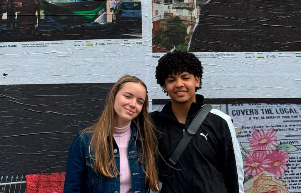 Fotografia de duas pessoas posando juntas e sorrindo diante de um mural com colagens de fotos e textos. À esquerda, uma pessoa de pele clara, cabelos lisos e loiros, usa jaqueta jeans sobre suéter rosa de gola alta. À direita, uma pessoa de pele parda, cabelos curtos e cacheados, veste jaqueta preta com detalhe branco e logotipo da Puma, além de uma bolsa a tiracolo. O fundo apresenta imagens impressas de paisagens urbanas e elementos gráficos coloridos.
