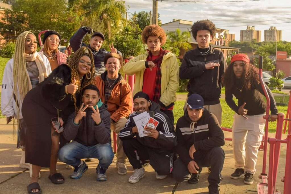 Fotografia colorida mostra um grupo diverso de 13 jovens negros e negras reunidos em uma área aberta com árvores e prédios ao fundo. Eles posam sorrindo e fazendo gestos com as mãos, expressando alegria, orgulho e união. Alguns usam roupas esportivas, outros casacos estilosos, gorros e tranças longas. Um dos jovens, agachado no centro, segura publicações com a capa visível, indicando participação em atividades culturais ou educativas. A luz do sol ilumina suavemente o grupo, criando uma atmosfera acolhedora e de pertencimento comunitário.